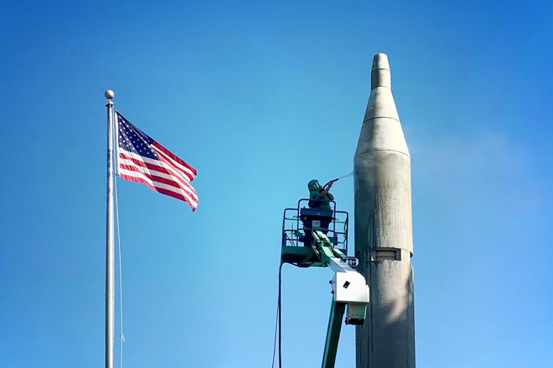 Abrasive Blaster on lift cleaning the top of a rocket with american flag in background